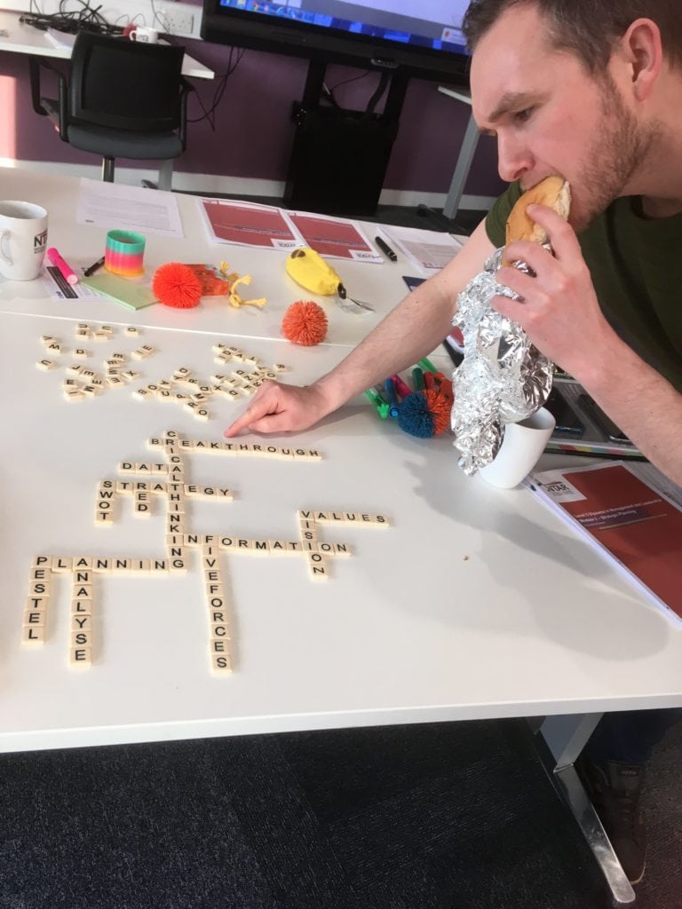One of our talent development programme members, Jonathan, eats a bacon barm while pointing to the word "breakthrough" written in scrabble letters